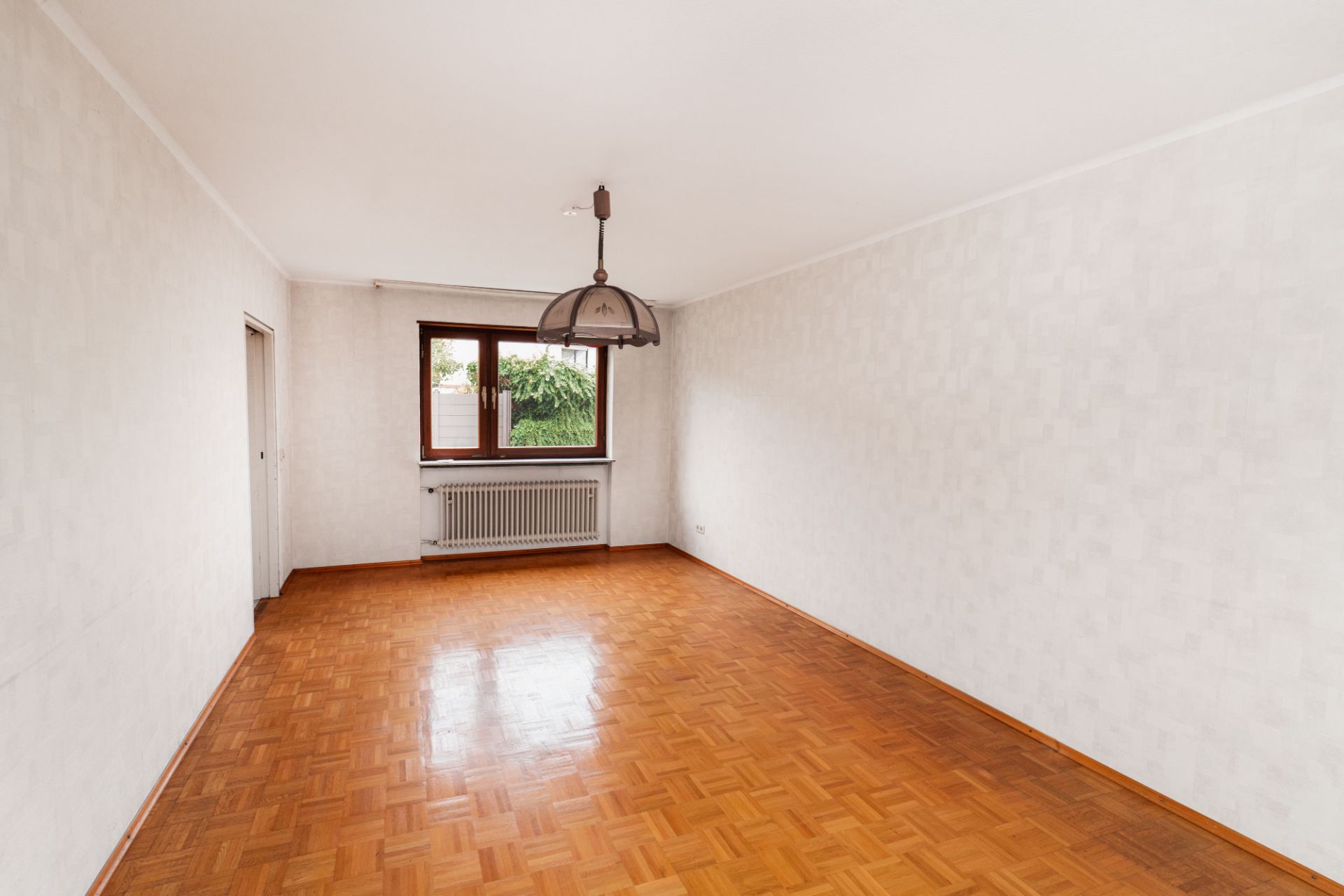 Leerer Raum mit Parkettboden, weiß gestrichenen Wänden, Fenster und Deckenlampe.