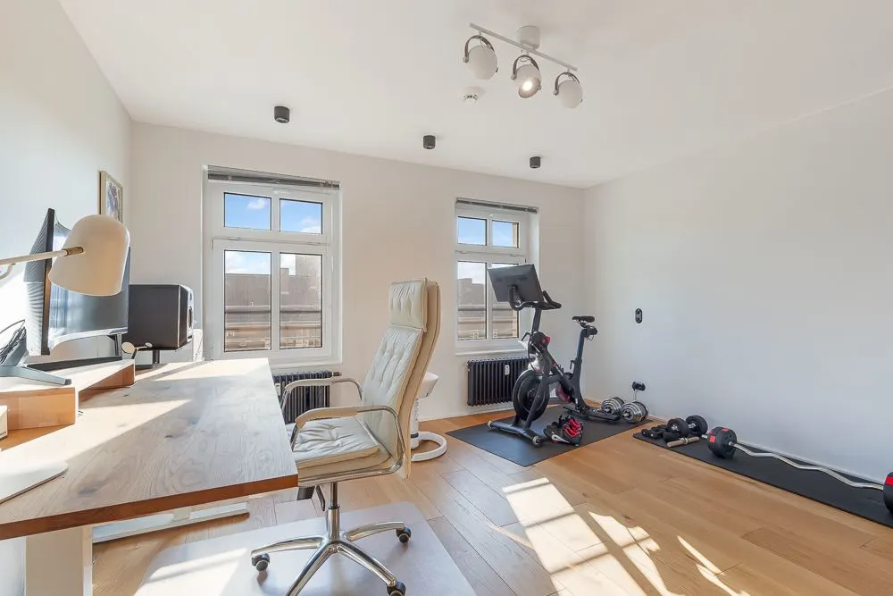 Helles Zimmer mit Schreibtisch, Bürostuhl, Heimtrainer und Hanteln auf Holzfußboden.