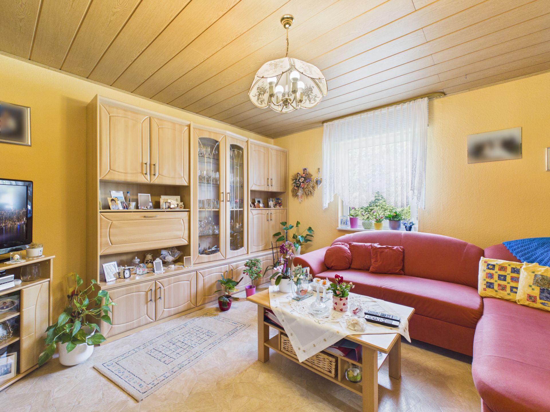 Wohnzimmer mit rotem Sofa, Holzmöbeln, Couchtisch und Pflanzen vor einem Fenster mit Gardine.