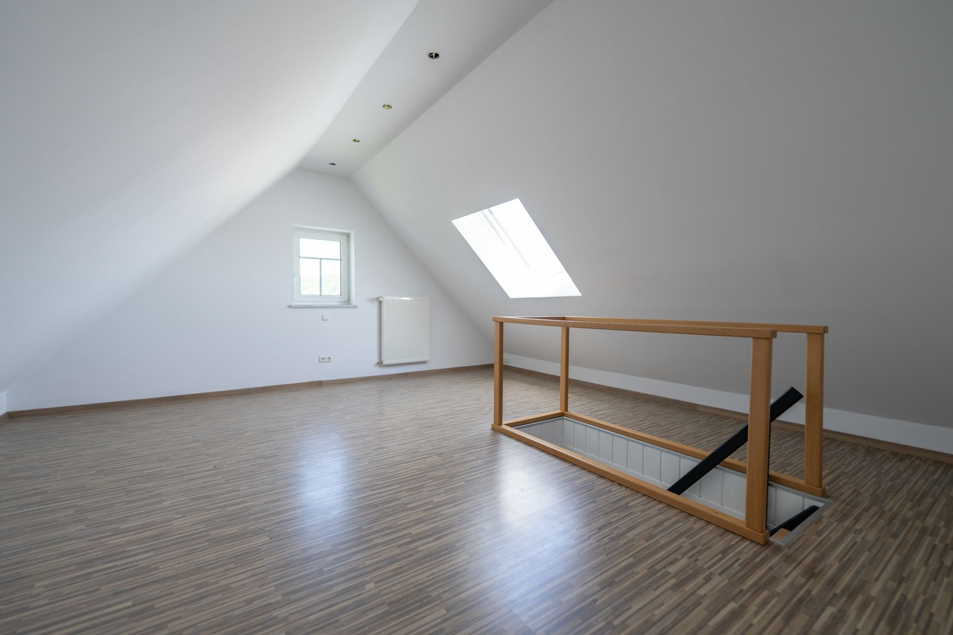 Leerer Dachraum mit Holzfußboden, Fenster, Dachfenster und Treppenöffnung mit Geländer.