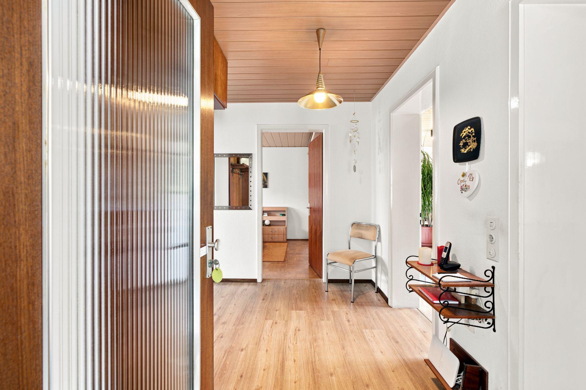 Heller Flur mit Holzboden, Stuhl, Wandregal, Wanddekoration und offener Tür zum Zimmer.