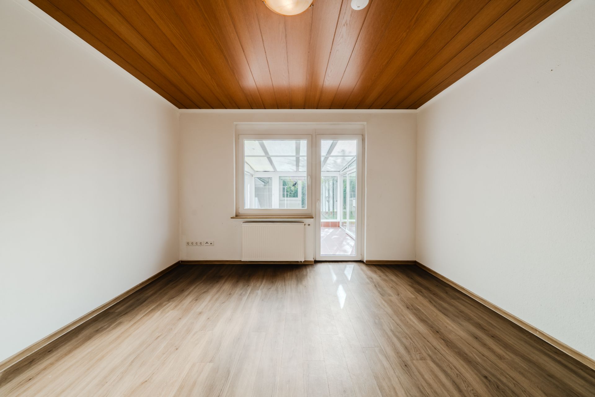 Leerer Raum mit Holzdecke, Holzfußboden, Fenster und Tür zu einem Wintergarten.