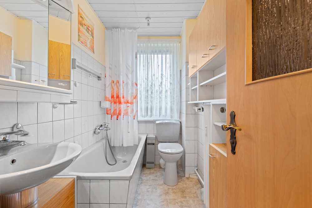 Helles Badezimmer mit Badewanne, Waschbecken, Toilette, Holzschränken und Fenster.