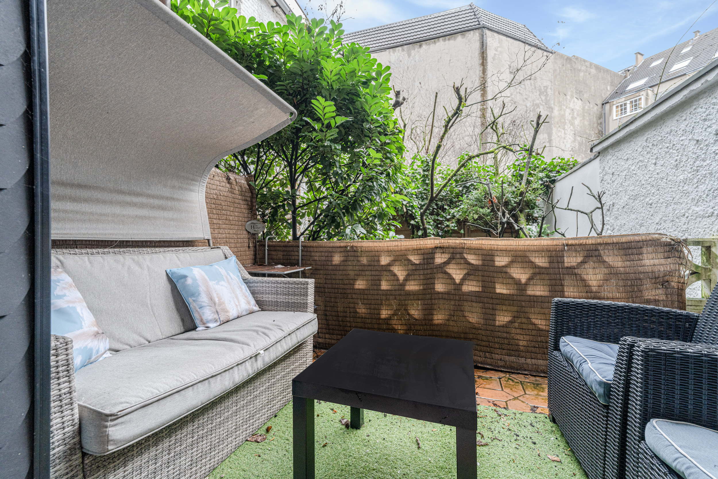 Kleine Terrasse mit Sofa, Sessel, Tisch und grünem Kunstrasen, umgeben von Pflanzen.