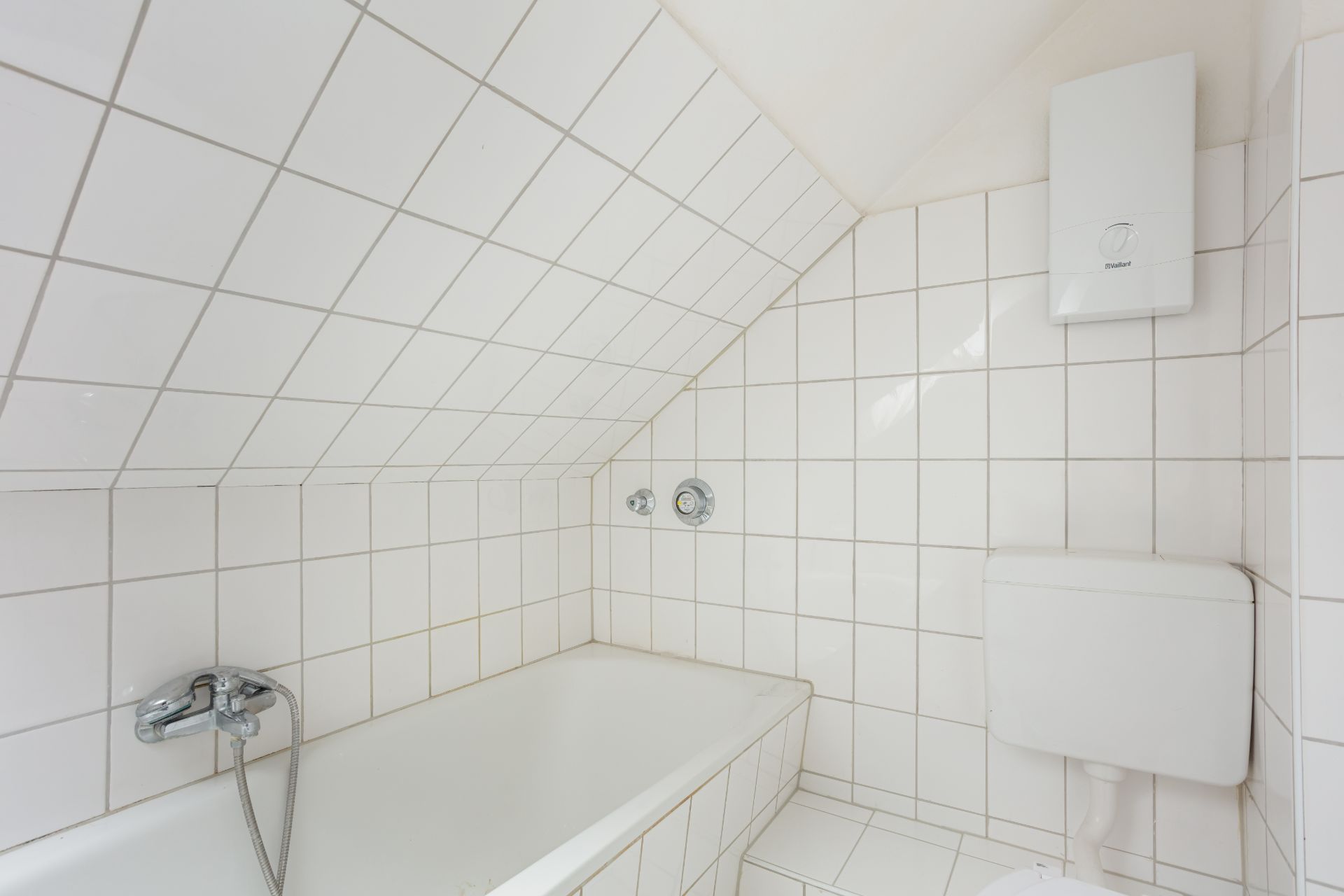 Weiß gefliestes Badezimmer mit Badewanne, Toilette und Wasserboiler unter Dachschräge.