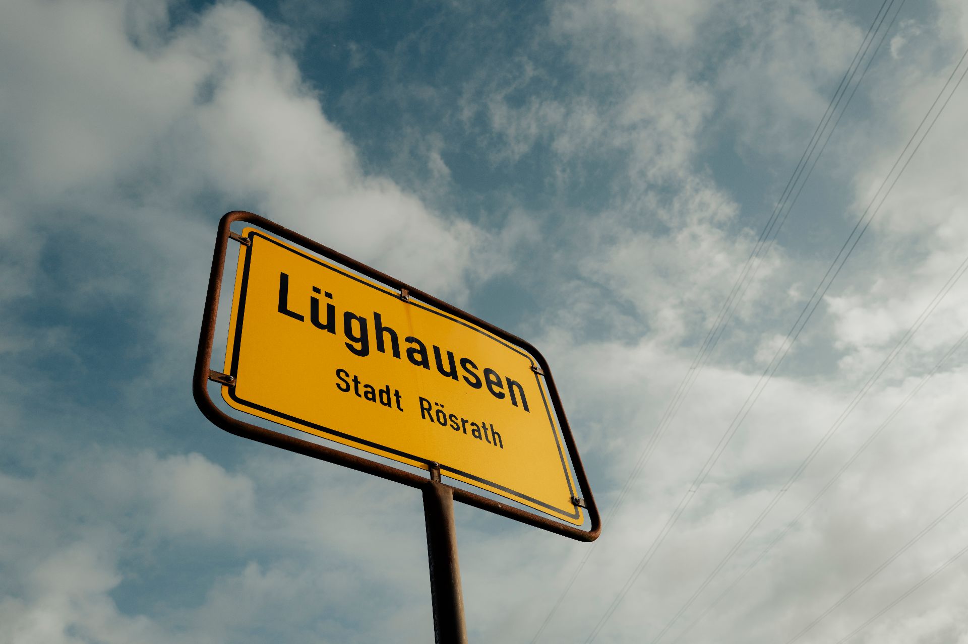 Gelbes Ortsschild mit der Aufschrift 'Lüghausen Stadt Rösrath' vor bewölktem Himmel.