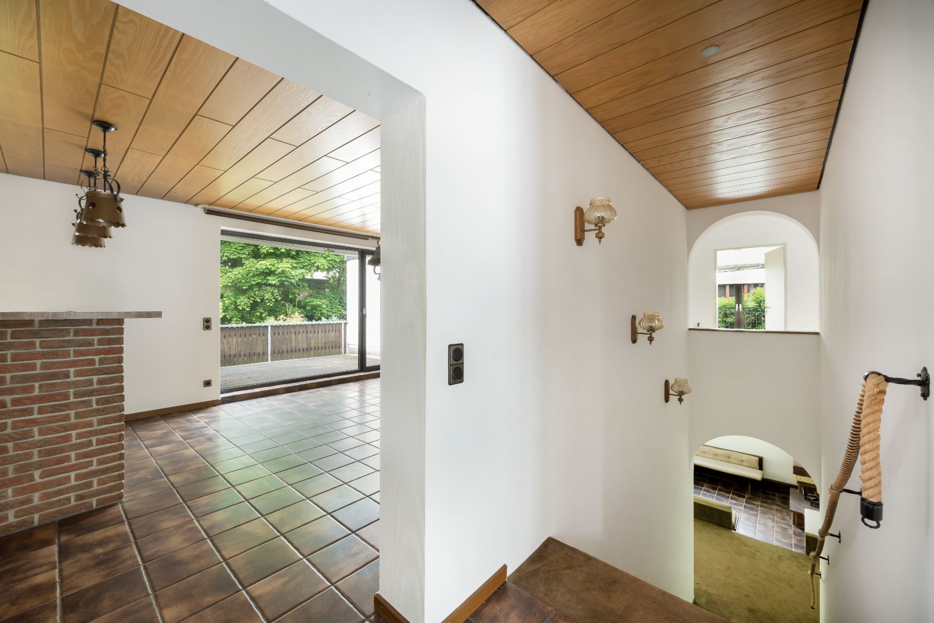 Flur mit Blick auf Wohnzimmer, große Fenster, Fliesenboden und Holzdecke, daneben Treppe nach unten.