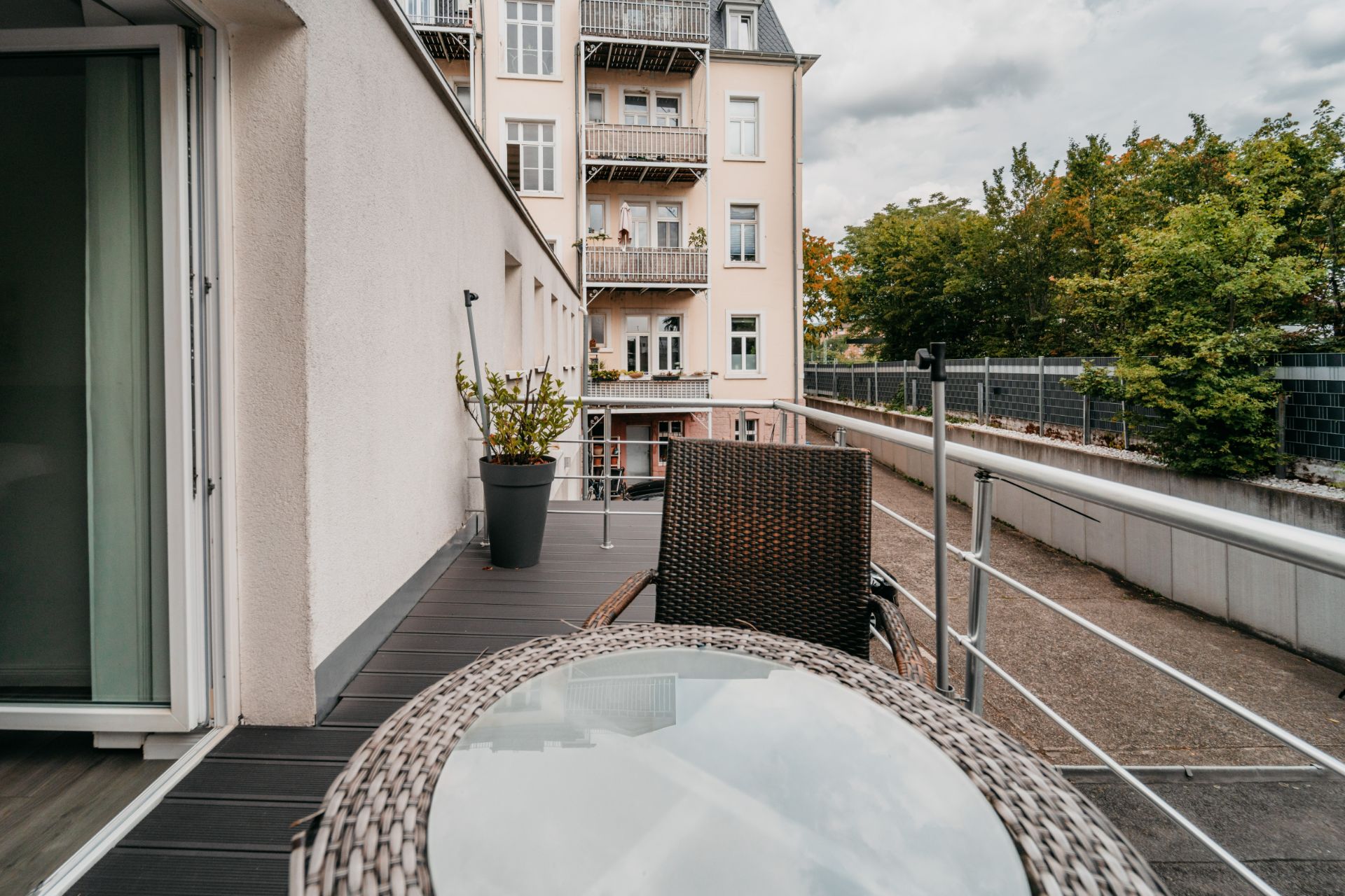 Balkon mit Glas-Tisch, Stuhl und Pflanze, Blick auf Nachbargebäude und Bäume.