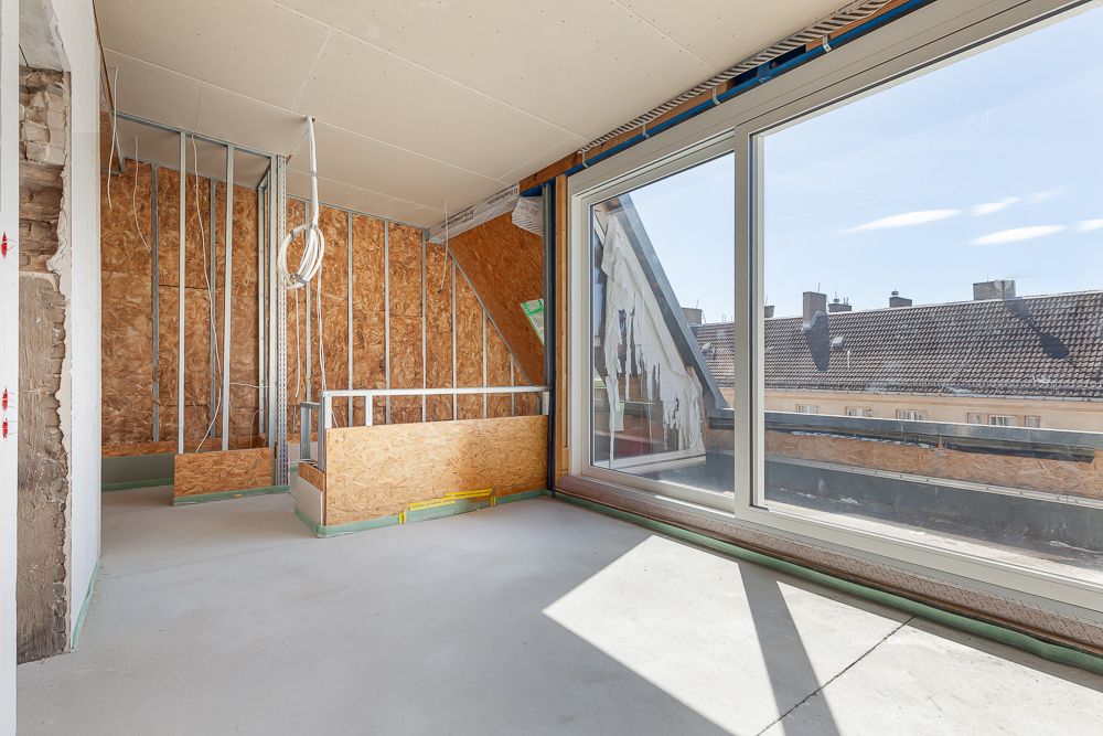 Helles Dachgeschosszimmer im Rohbau mit großen Fenstern und Blick auf Dächer.