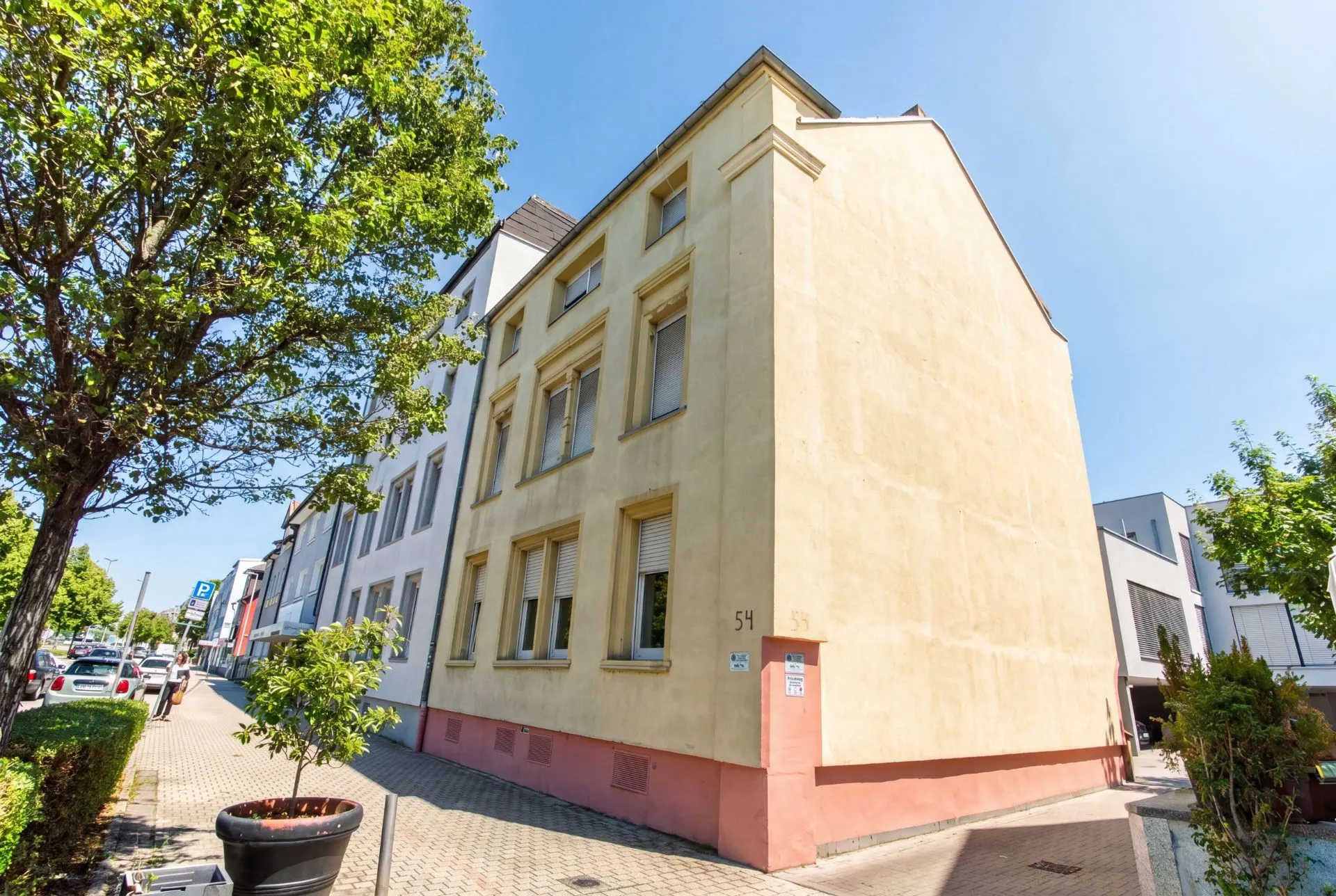 Gelbes Wohnhaus mit mehreren Fenstern an einer Straße mit Bäumen und geparkten Autos.