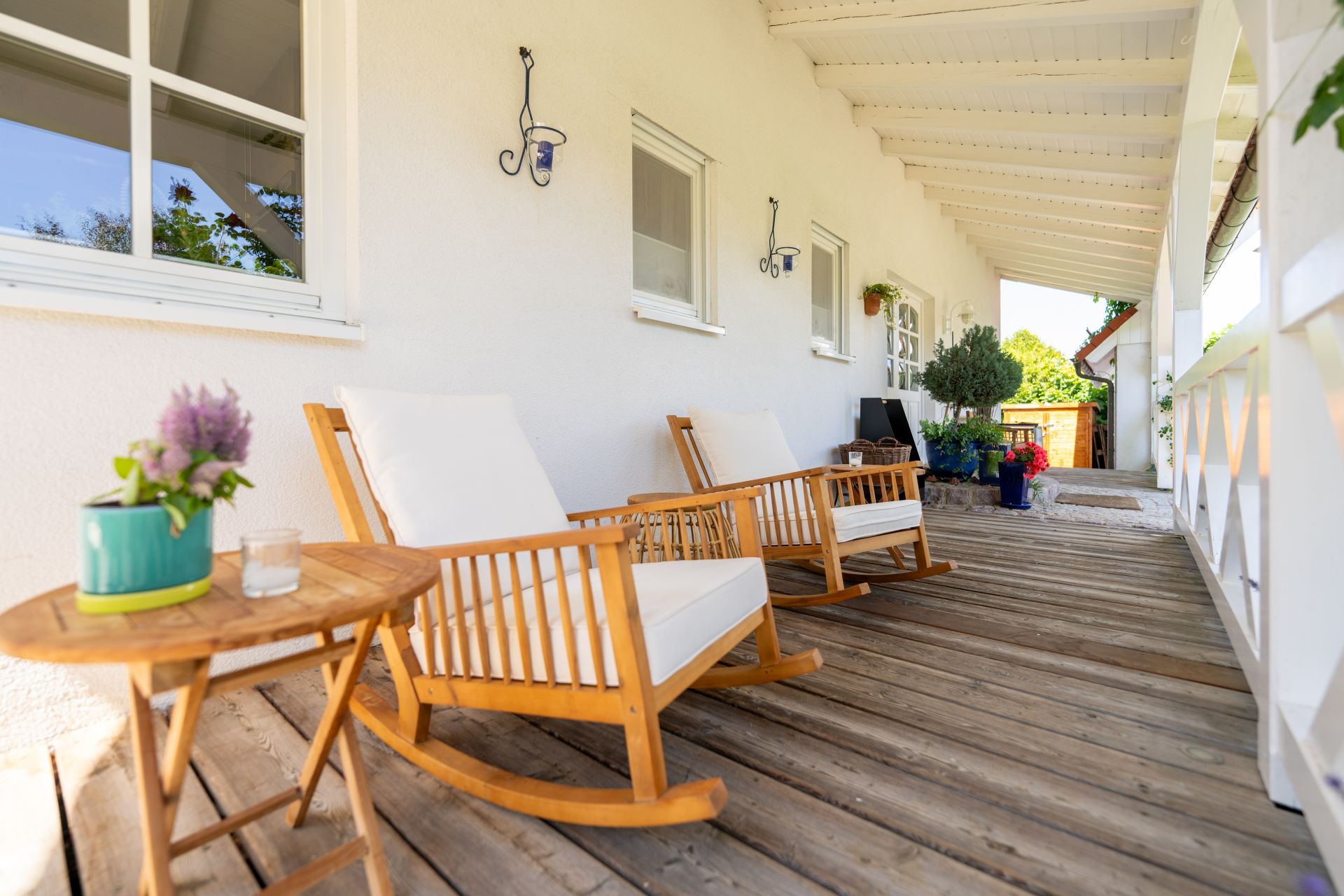 Holzveranda mit zwei weißen Schaukelstühlen, kleinem Tisch und Pflanzen an einer weißen Hauswand.