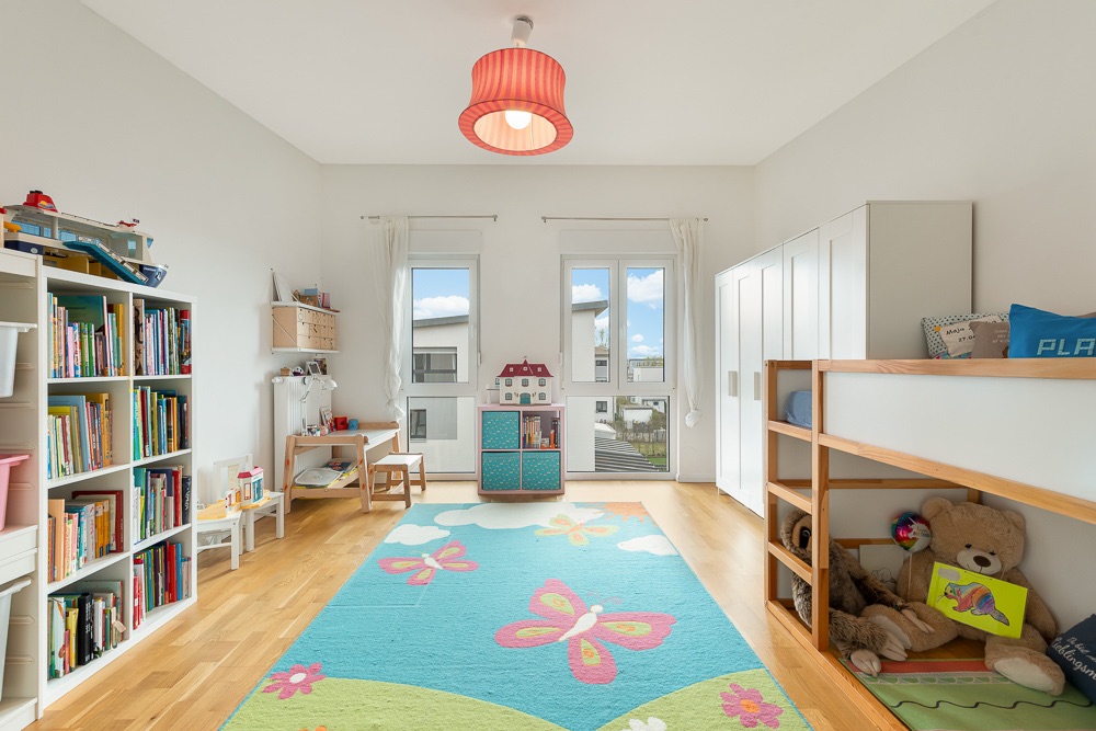 Helles Kinderzimmer mit Hochbett, Bücherregal, Spielzeug und buntem Teppich mit Schmetterlingen.