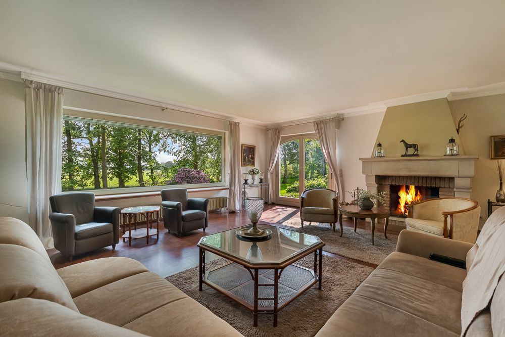 Wohnzimmer mit großen Fenstern, Kamin, mehreren Sesseln und Sofas sowie Blick ins Grüne.