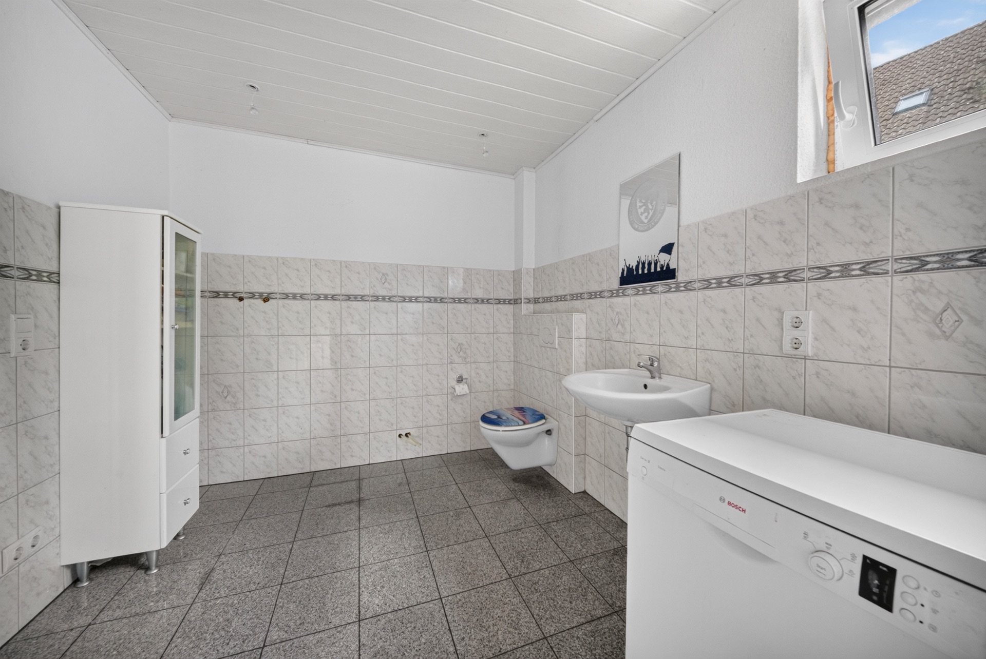 Heller, gefliester Raum mit Waschbecken, Wandtoilette, weißem Schrank und Waschmaschine unter Fenster.