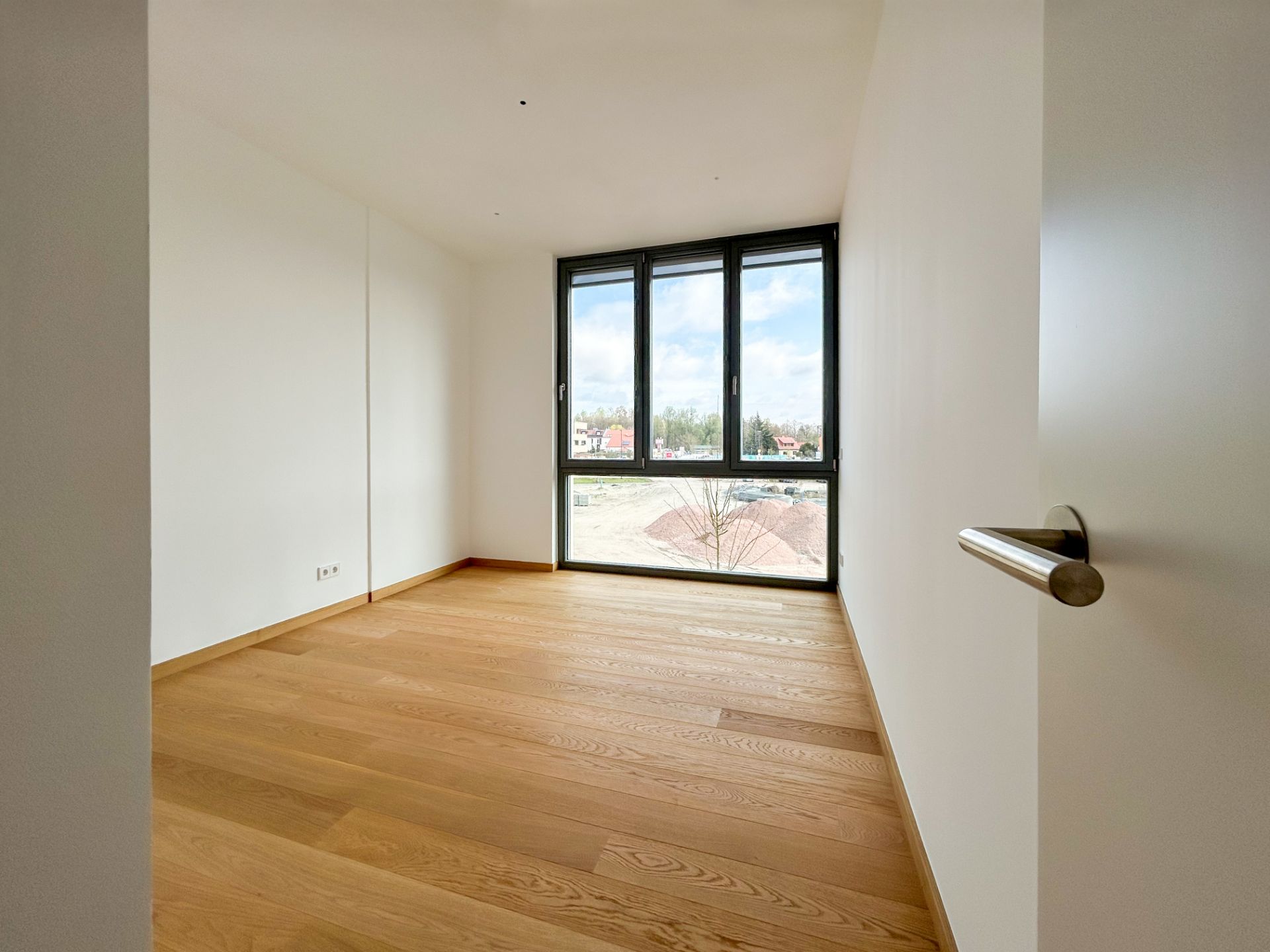 Helles, leeres Zimmer mit Holzfußboden und großem Fenster mit Blick nach draußen.