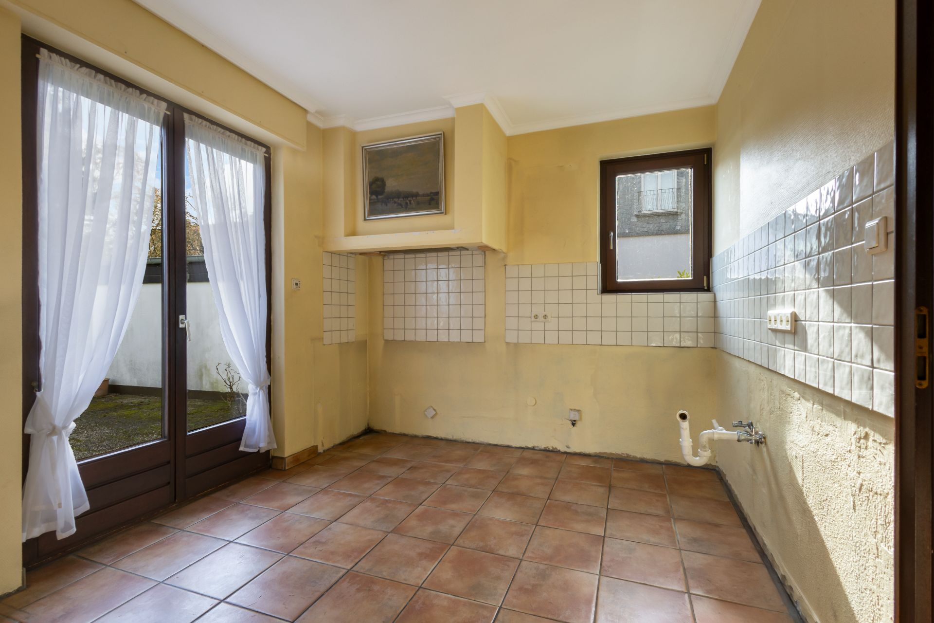 Leerer Raum mit Fliesenboden, gefliester Wand, Fenster und Glastür mit weißen Vorhängen.