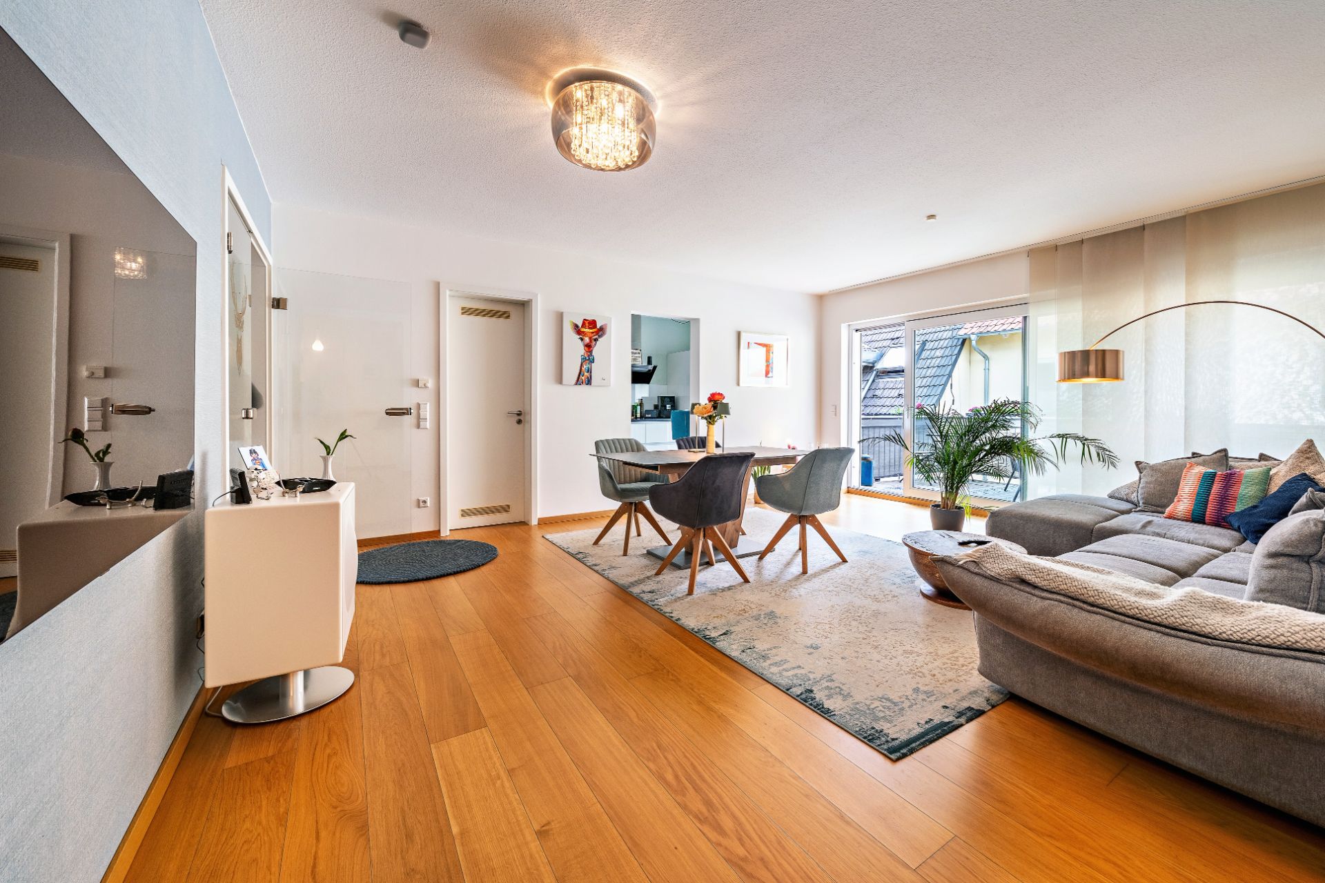 Helles Wohnzimmer mit grauem Sofa, Esstisch, Holzfußboden und großen Fenstern zur Terrasse.