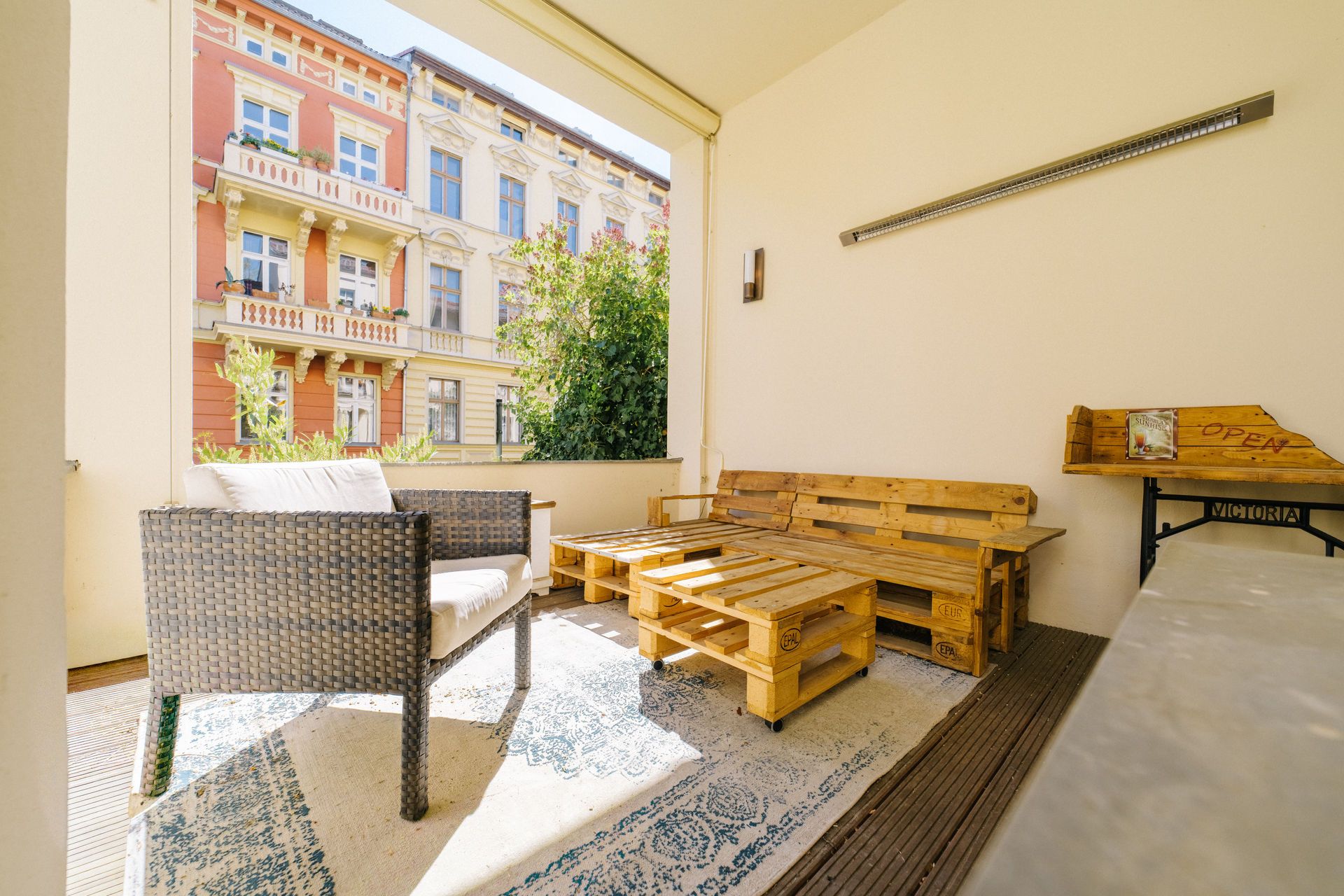 Überdachter Balkon mit Sitzmöbeln aus Holzpaletten und einem Sessel, Blick auf Nachbargebäude.