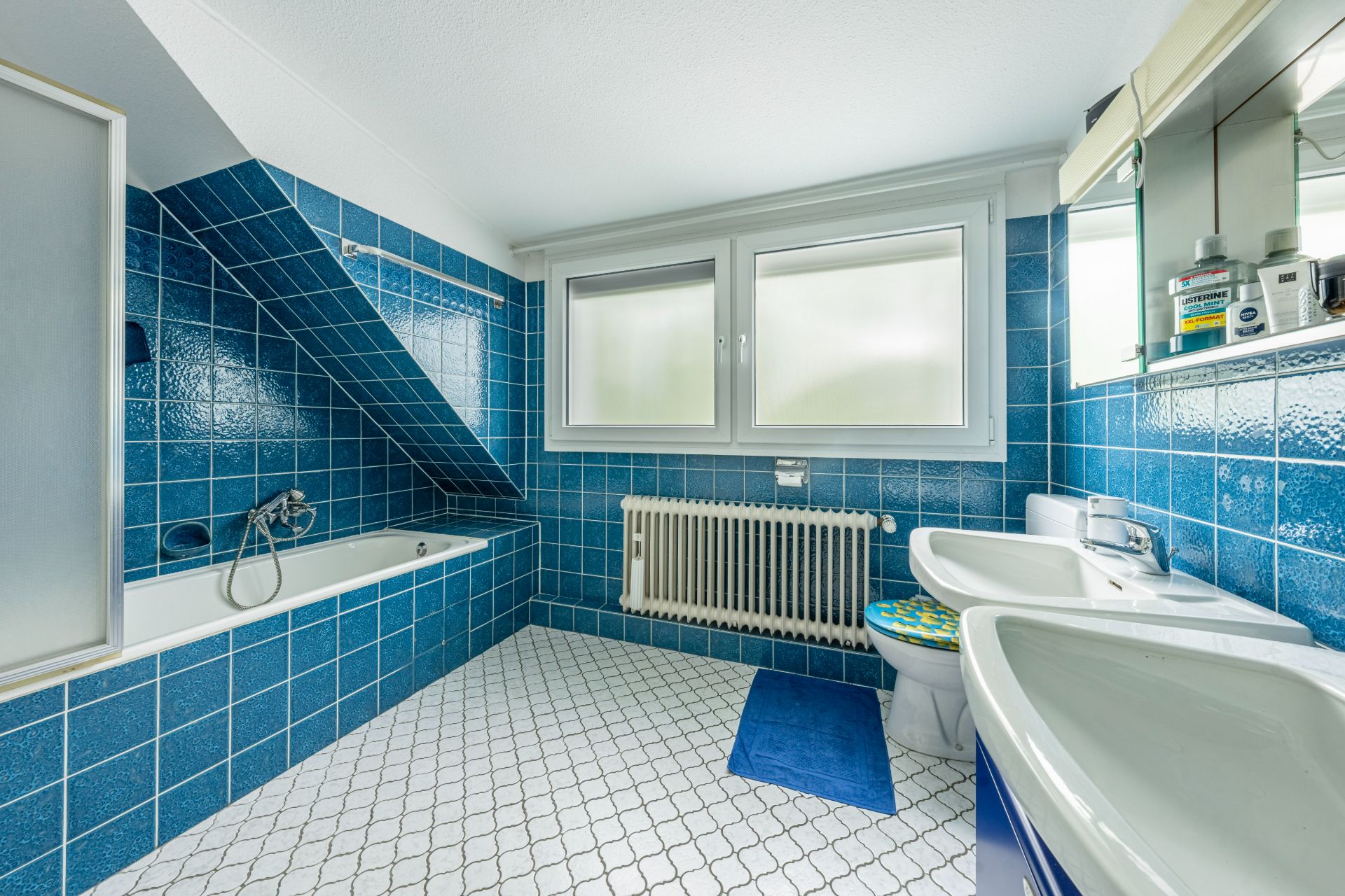 Badezimmer mit blauen Fliesen, Badewanne, zwei Waschbecken und zwei Fenstern.