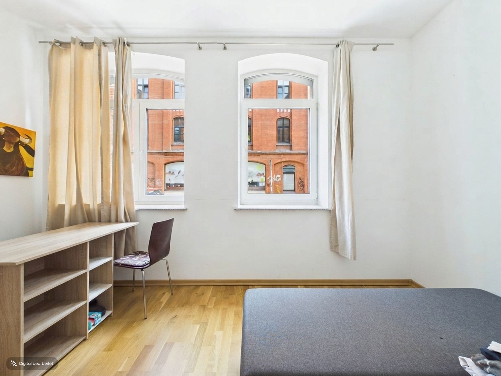 Helles Zimmer mit zwei Fenstern, einem Schreibtisch, Stuhl, Regal und Teil eines Bettes.
