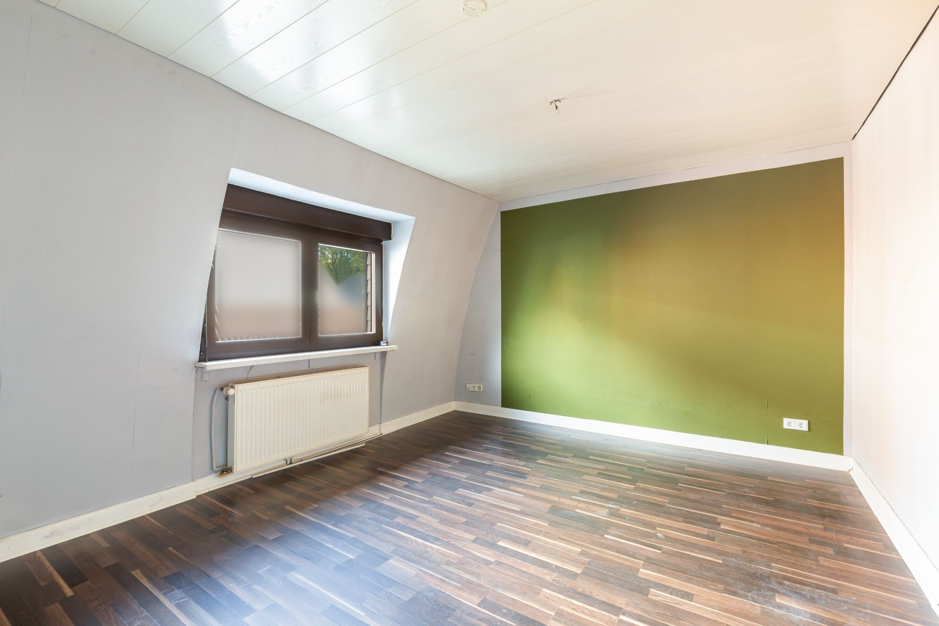 Leerer Raum mit dunklem Holzboden, grün gestrichener Wand und Fenster mit Heizung darunter.