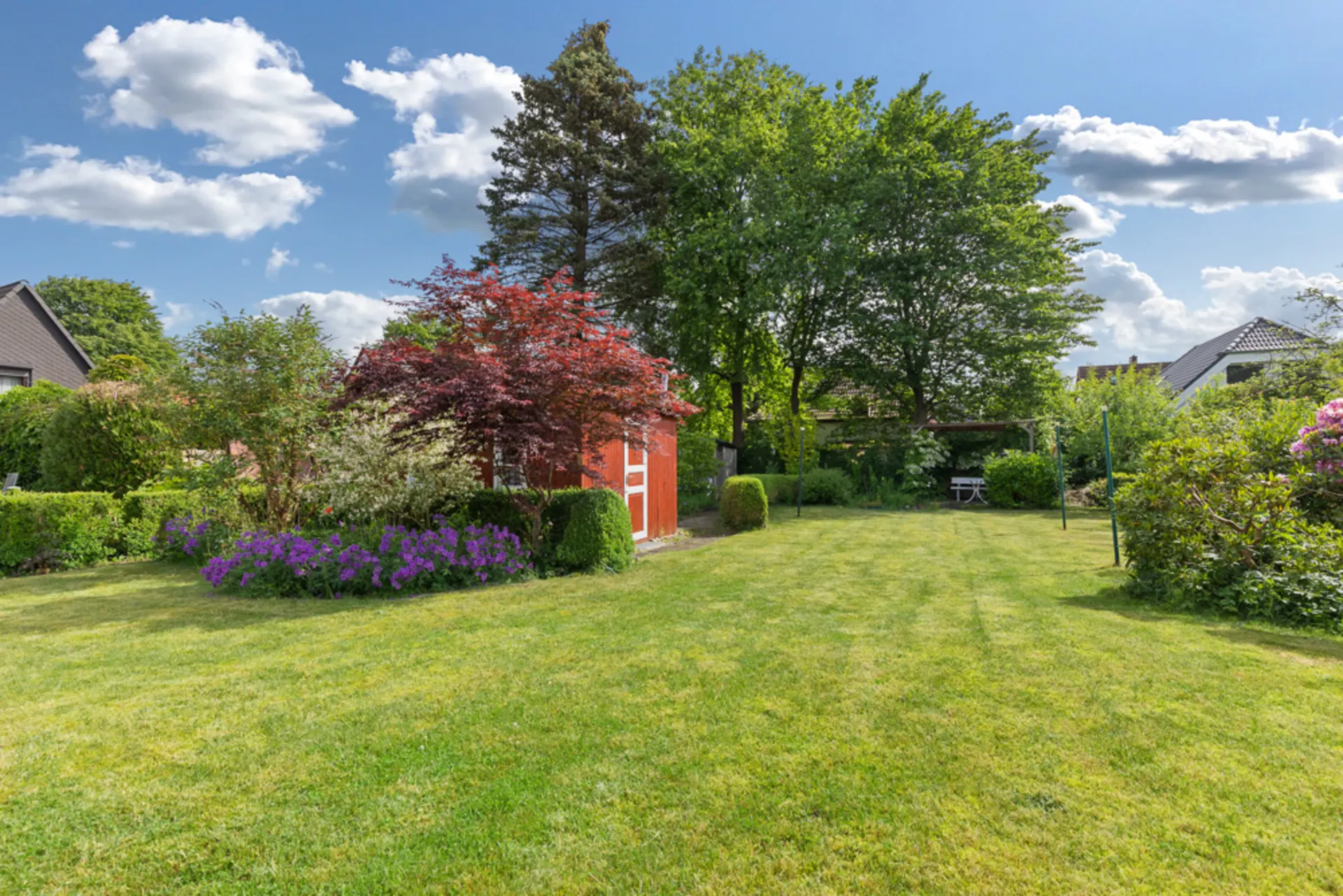 Großer Garten mit Rasen, Blumenbeet, rotem Gartenschuppen und Bäumen bei sonnigem Wetter.