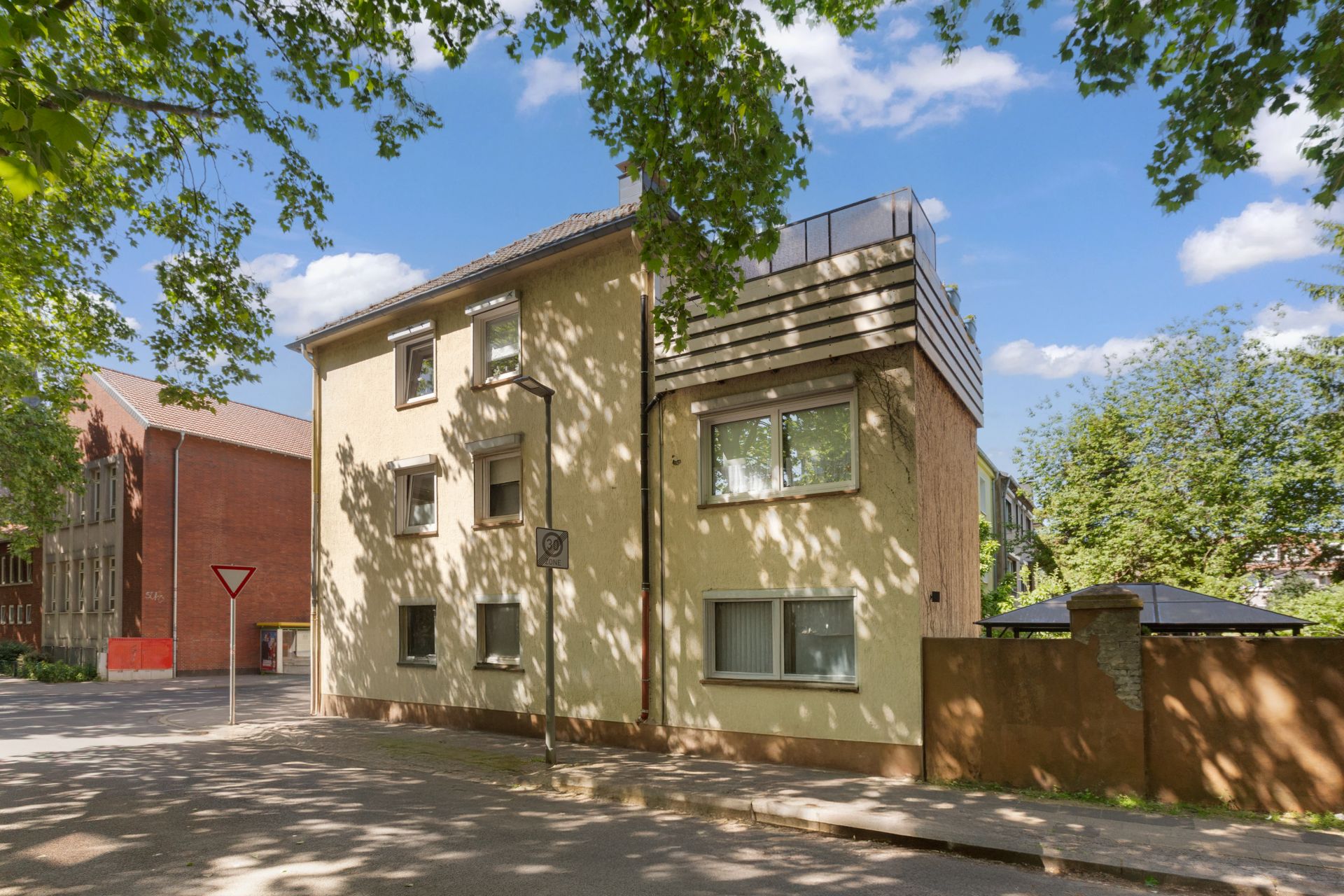 Beiges Wohnhaus mit mehreren Fenstern an einer ruhigen Straße, Bäume spenden Schatten.