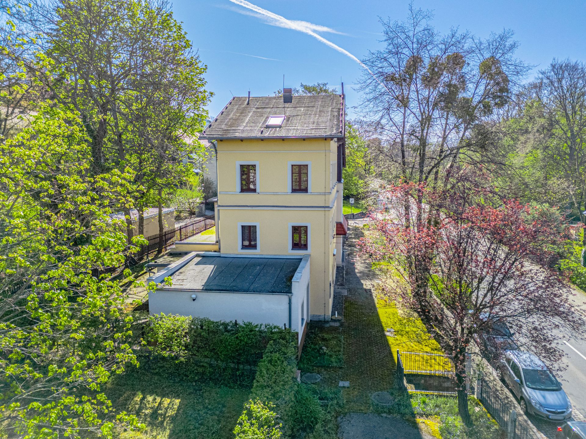Gelbes Wohnhaus mit Garten und Bäumen an einer Straße bei sonnigem Wetter.