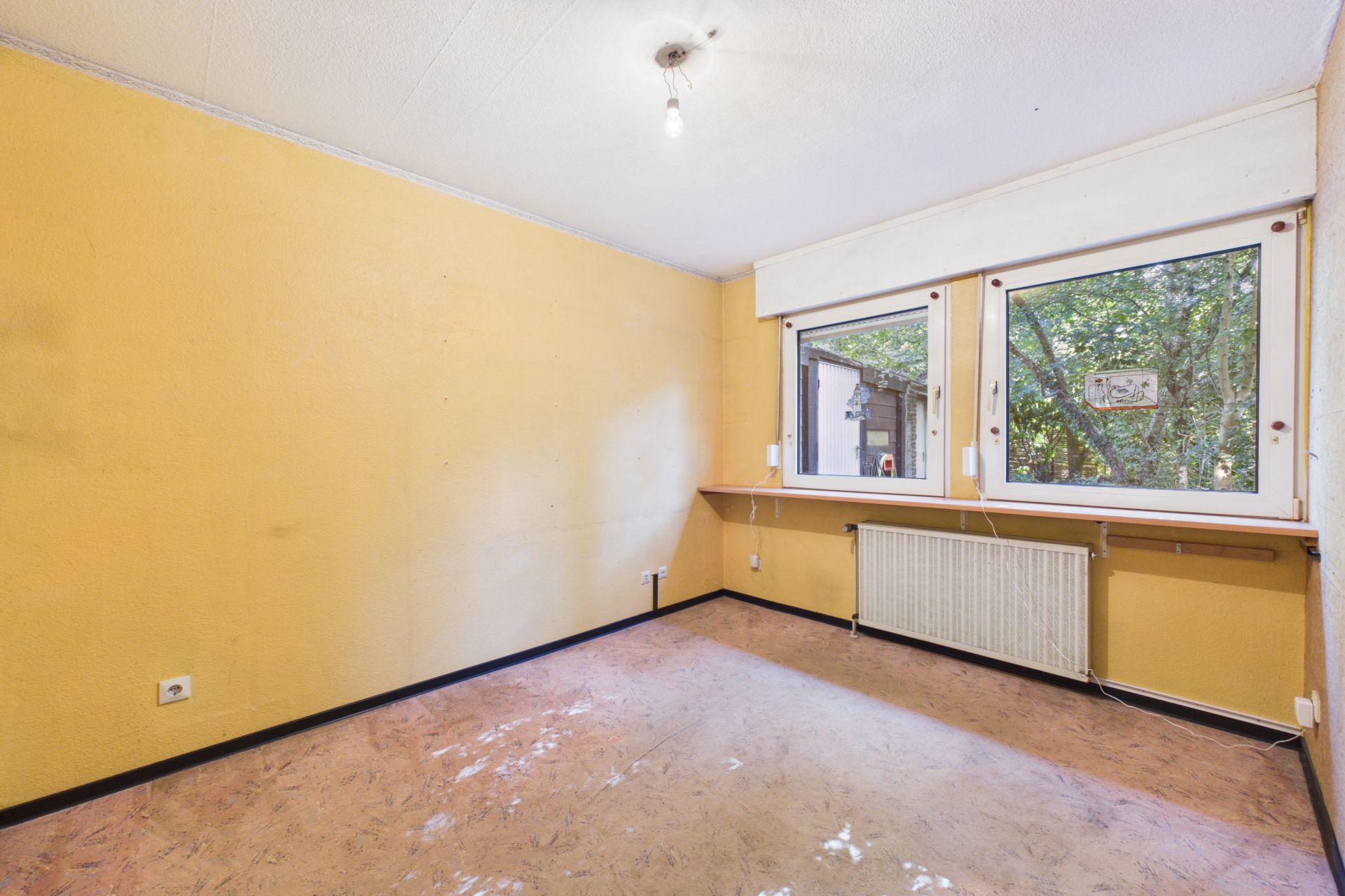 Leeres Zimmer mit gelben Wänden, zwei Fenstern, Heizung und einfacher Beleuchtung.