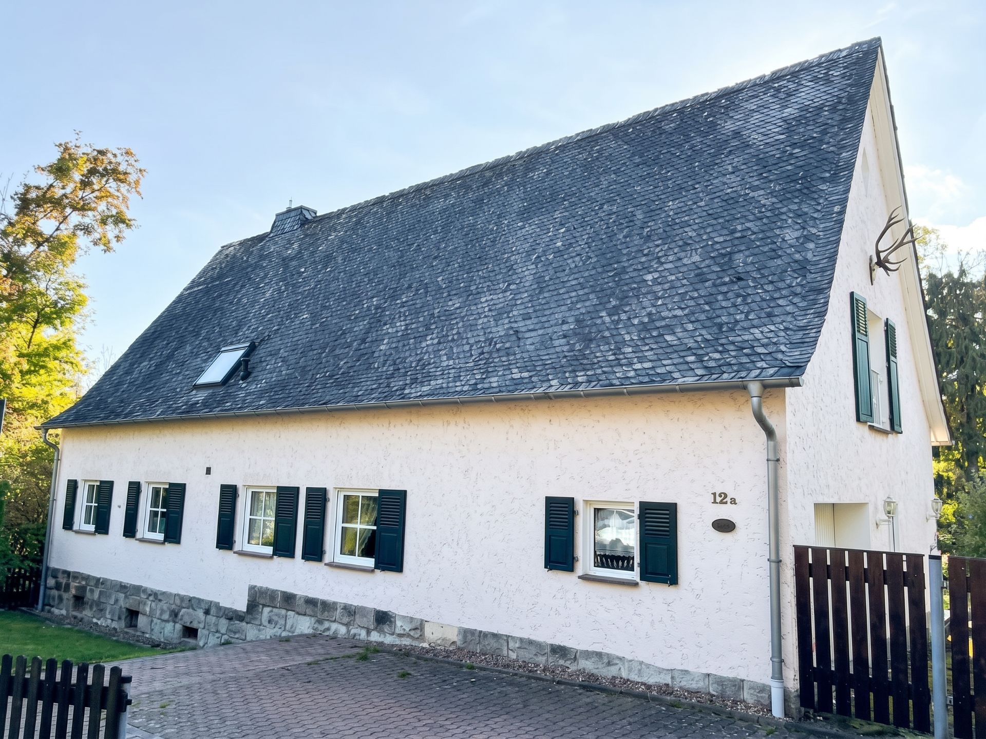 Weiße Hausfassade mit schwarzem Schieferdach, mehreren Fenstern und grünem Fensterläden.