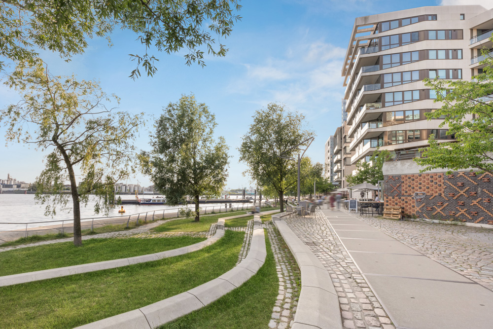 Moderne Wohngebäude am Flussufer mit Bäumen, Gehweg und Sitzbereichen im Grünen.