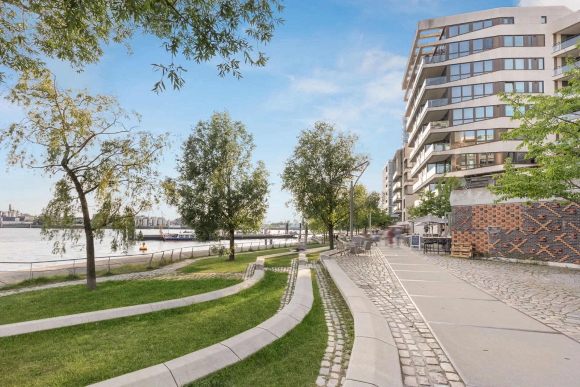Moderne Wohngebäude am Flussufer mit Bäumen, Gehweg und Sitzbereichen im Grünen.