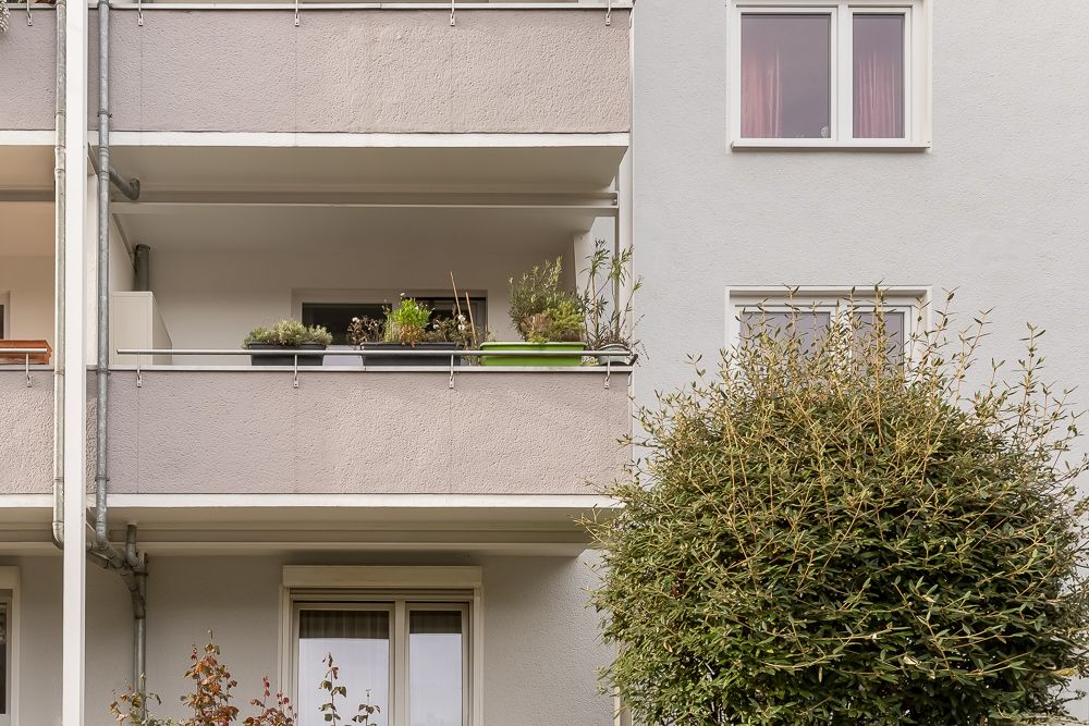 Mehrfamilienhaus mit Balkon und mehreren Pflanzkästen, daneben ein grüner Strauch.