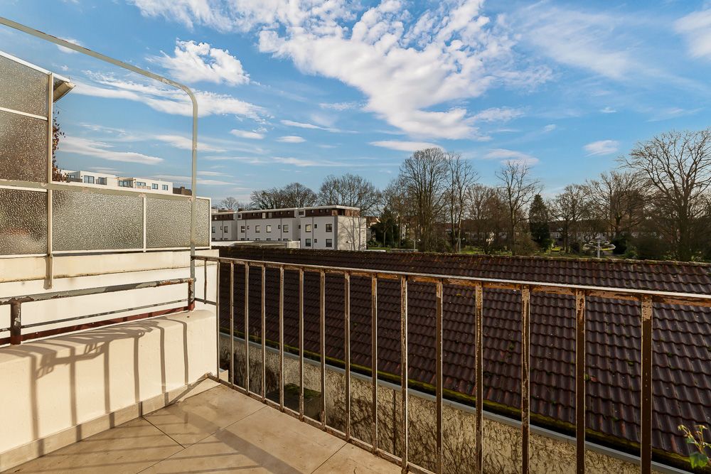 Balkon mit Metallgeländer, Blick auf Dächer, Bäume und Wohngebäude bei blauem Himmel.