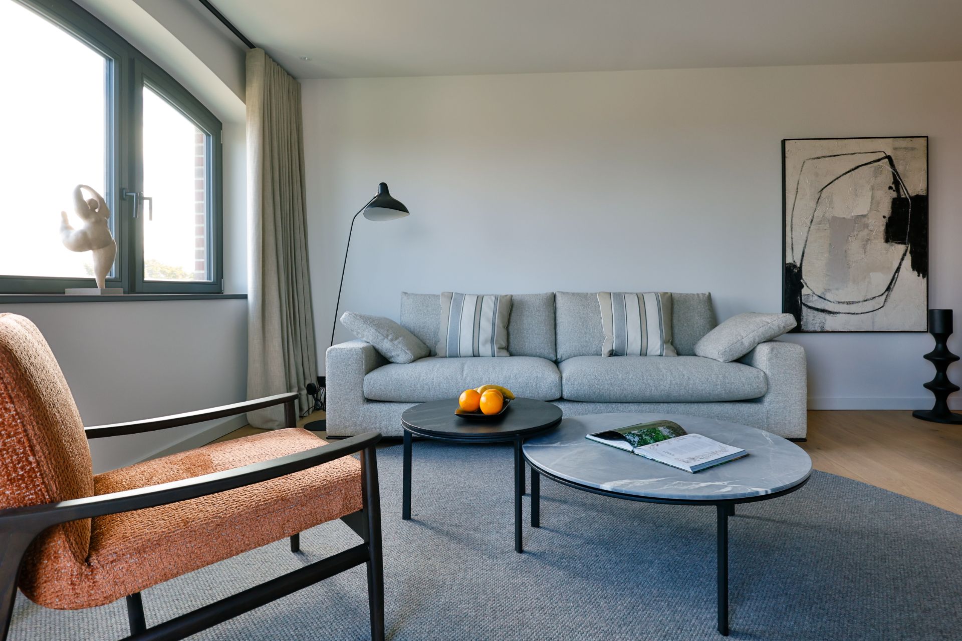 Helles Wohnzimmer mit grauem Sofa, zwei runden Tischen, Sessel und großem Fenster.