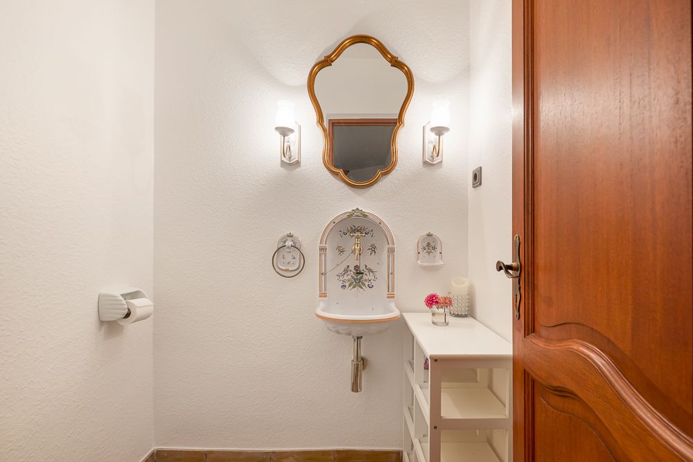 Kleines Badezimmer mit Waschbecken, Spiegel, Holzregal und goldenen Wandlampen.