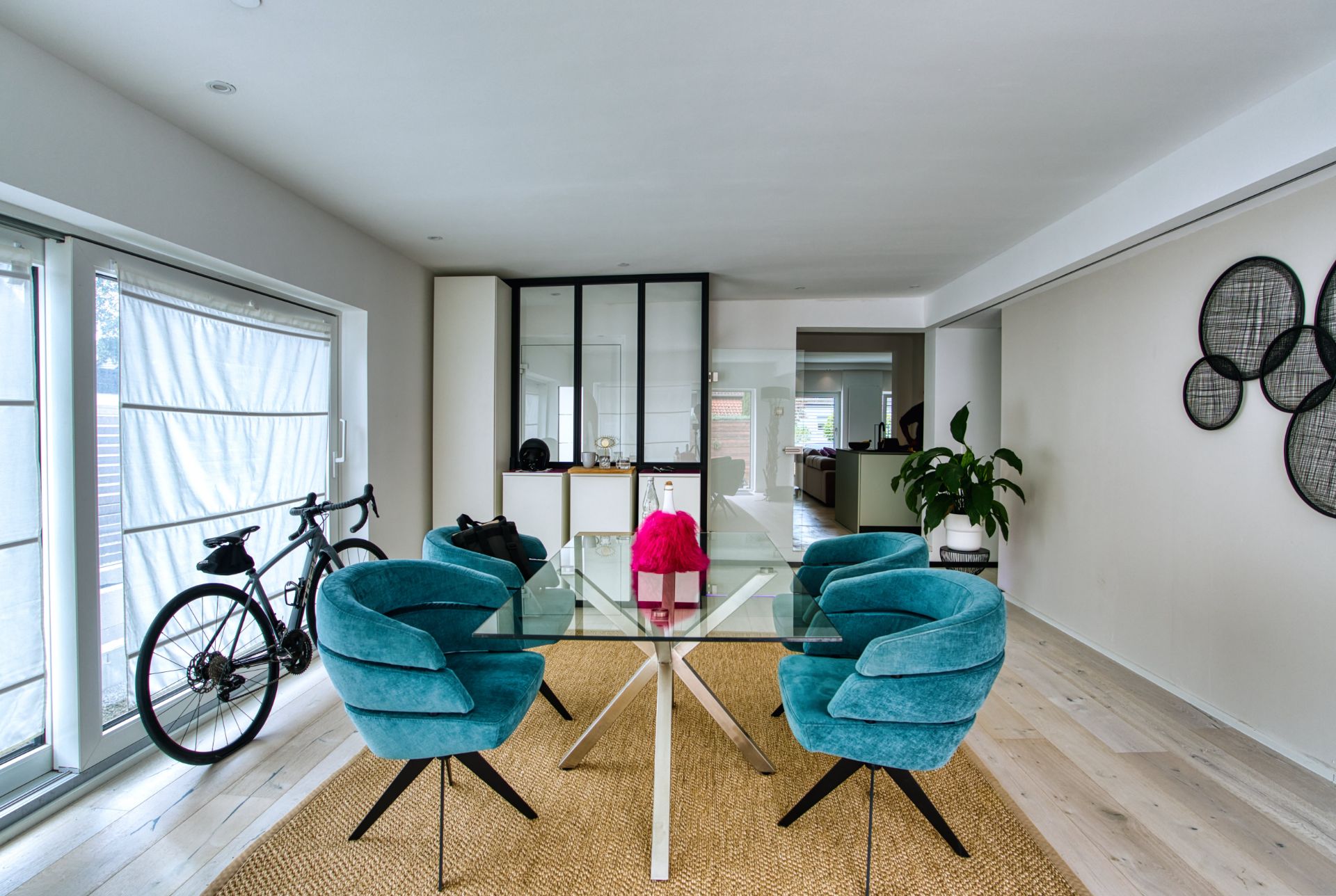 Esszimmer mit Glastisch, vier blauen Stühlen, Fahrrad und großer Fensterfront.