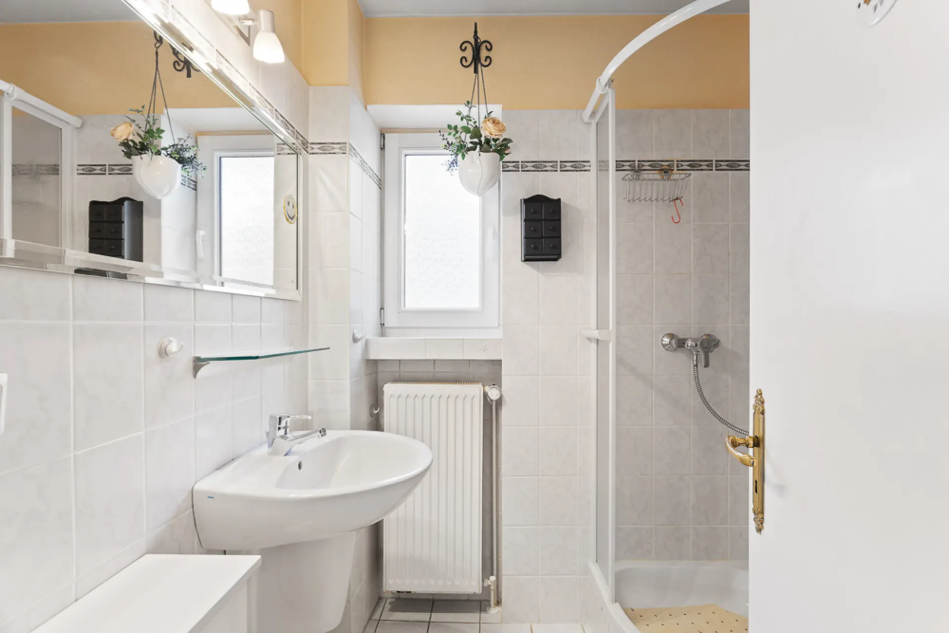Helles Badezimmer mit Waschbecken, Spiegel, Dusche, Fenster und Heizkörper.