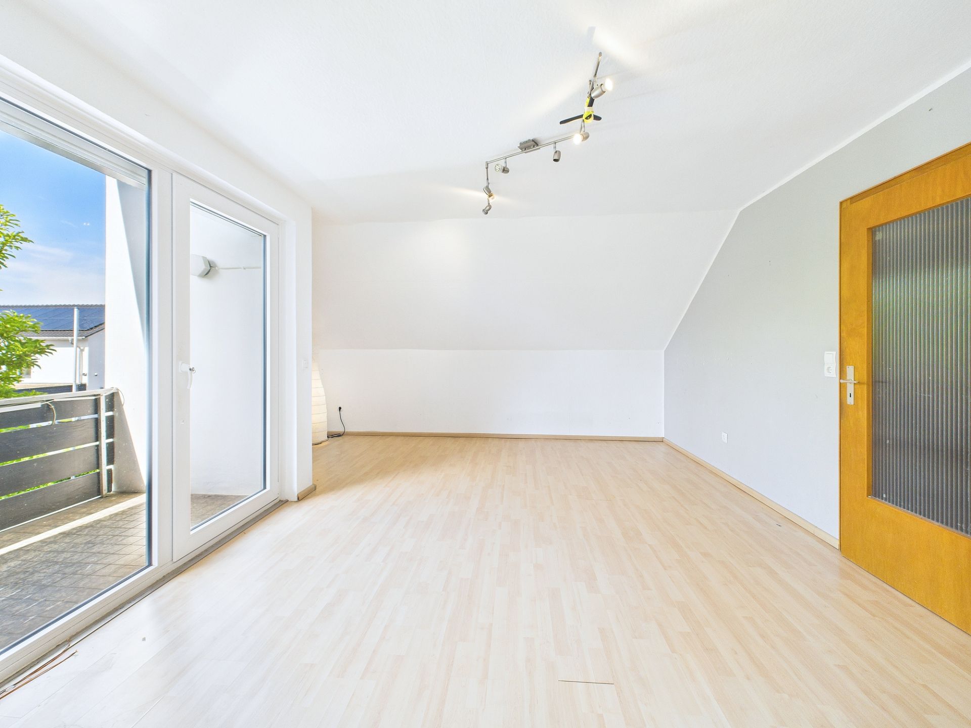 Helles, leeres Zimmer mit Holzfußboden, großer Fensterfront und Zugang zum Balkon.