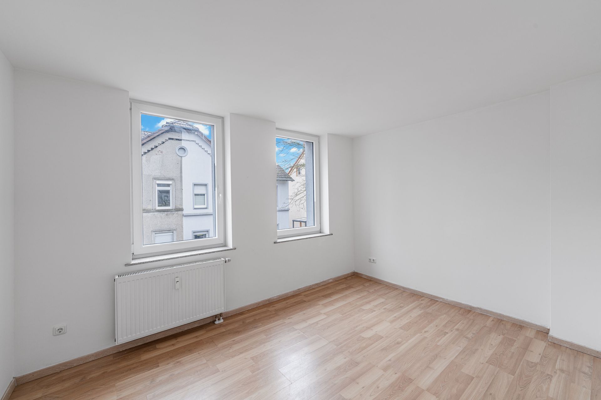 Leerer Raum mit hellem Laminatboden, zwei Fenstern und weiß gestrichenen Wänden.