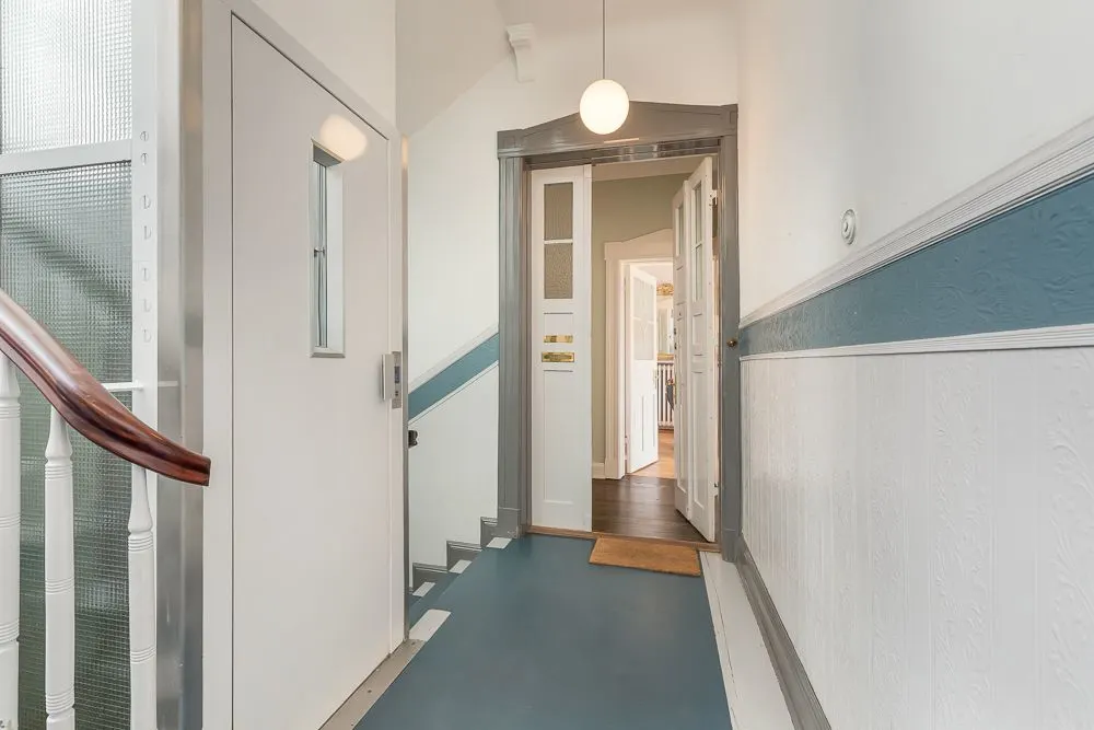 Flur mit Aufzugstür, Treppengeländer und geöffneter Tür zu einer Wohnung.