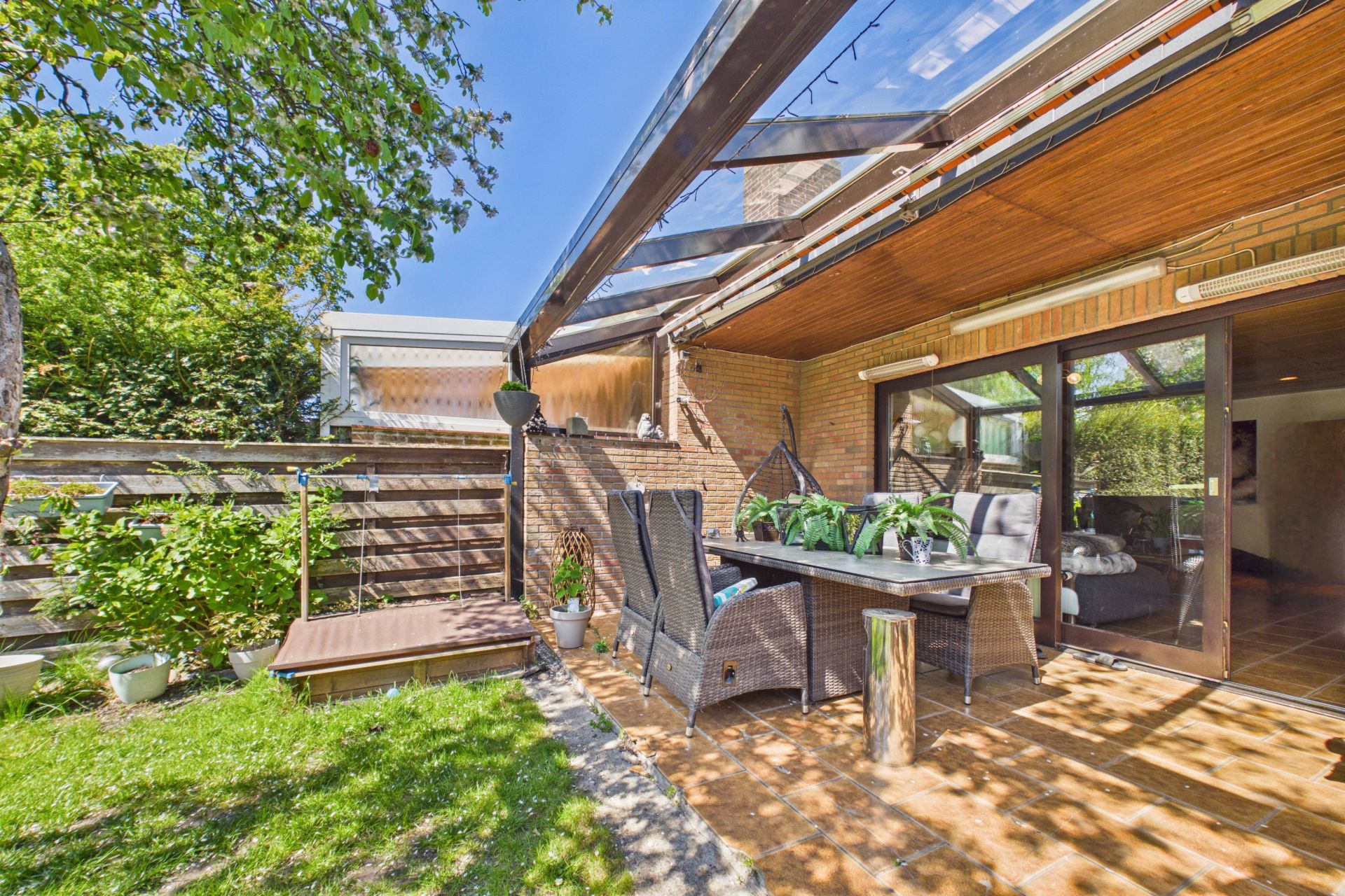 Überdachte Terrasse mit Gartentisch und Stühlen neben einer Rasenfläche, große Glastüren im Hintergrund.