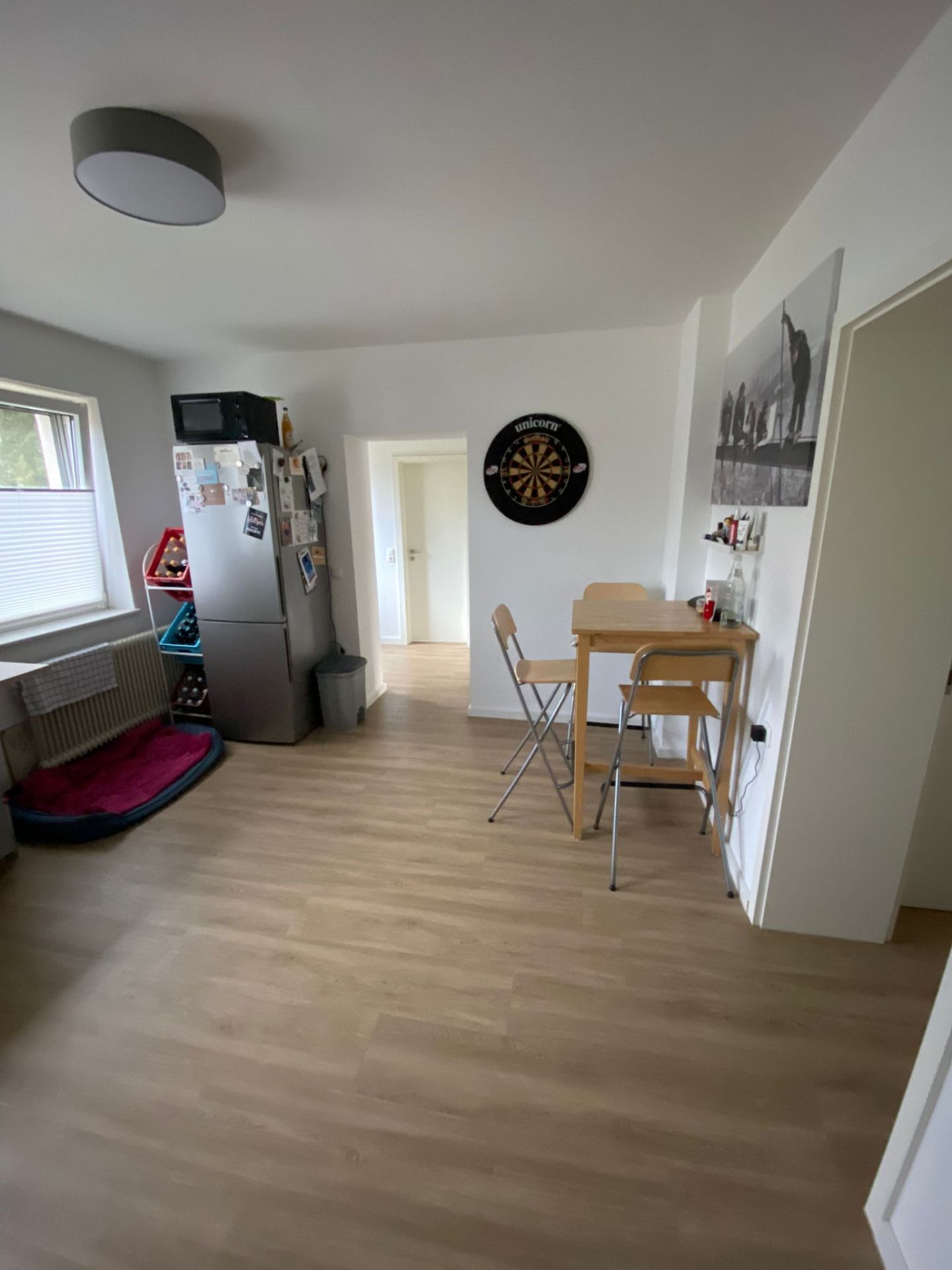 Wohnzimmer mit Tisch, zwei Stühlen, Kühlschrank, Fenster und Dartscheibe an der Wand.