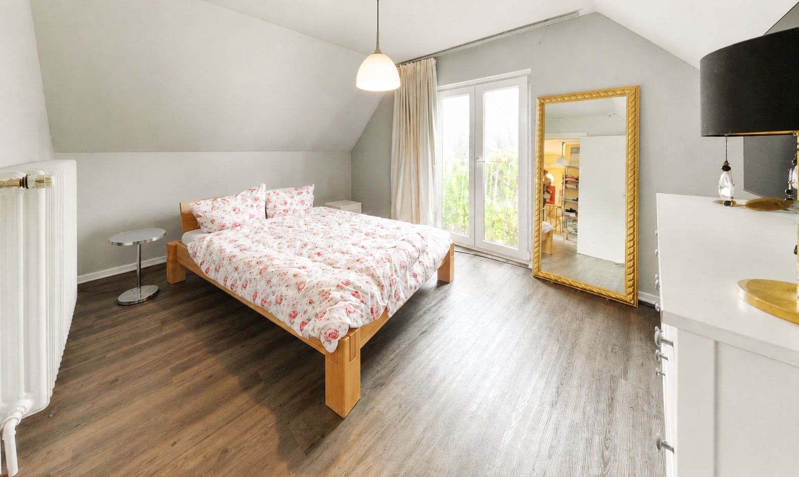 Helles Schlafzimmer mit Doppelbett, großem Spiegel, Holzboden und Fenstertüren zum Balkon.