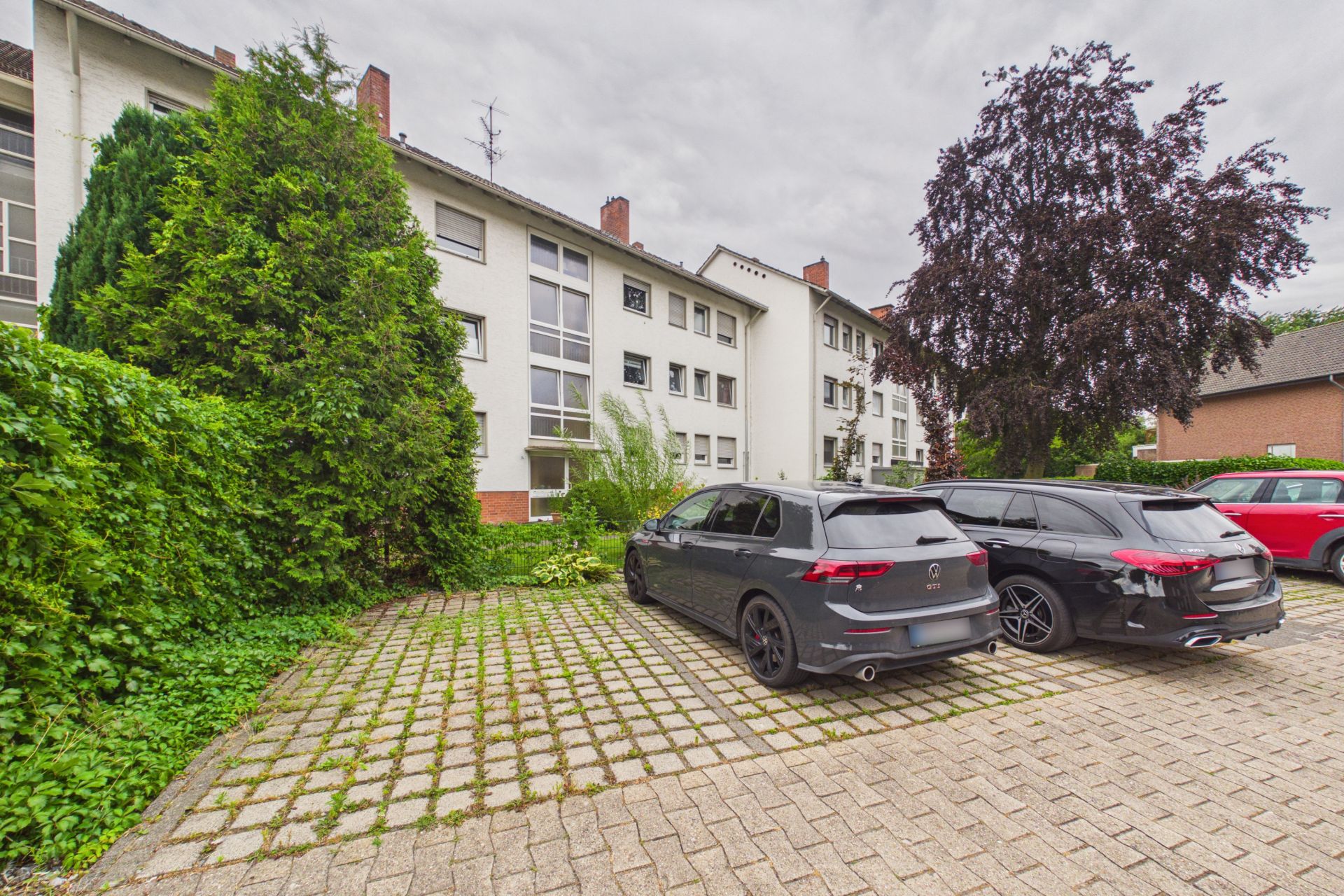 Mehrfamilienhaus mit Parkplatz, auf dem zwei Autos stehen, umgeben von Bäumen und Büschen.