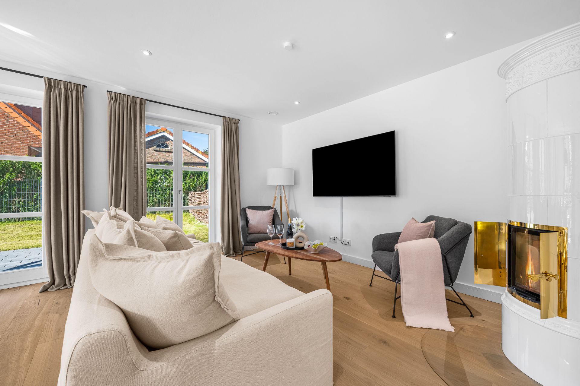 Helles Wohnzimmer mit Sofa, Sessel, Couchtisch, großem Fernseher und Kamin, Blick in den Garten.