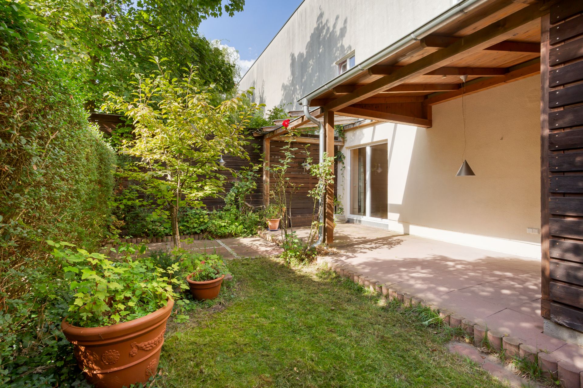 Kleiner Garten mit Terrasse, Topfpflanzen und Holzüberdachung an einem Wohnhaus.
