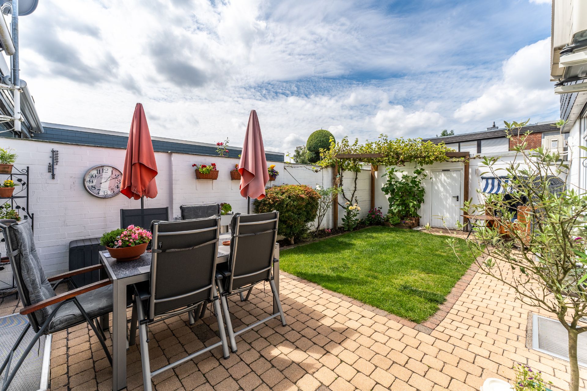 Gepflegter Garten mit Rasen, Terrasse, Gartentisch, Stühlen und zwei roten Sonnenschirmen.