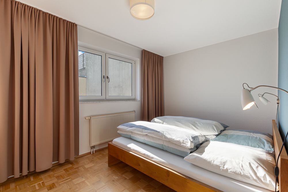 Helles Schlafzimmer mit Doppelbett, Parkettboden, Fenstern und braunen Vorhängen.