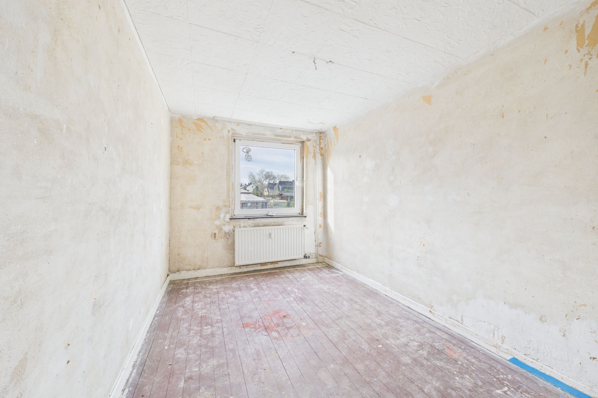 Leerer, renovierungsbedürftiger Raum mit abgenutztem Boden und Fenster an der Stirnwand.